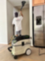 Person in white T-shirt sanding a ceiling with a Festool device. A silver fridge is visible. The setting is indoors and appears under renovation.