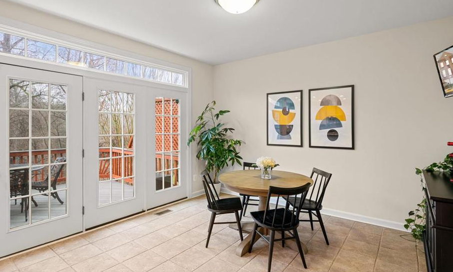Dining area with round table, four chairs, and a vase of flowers. Large windows show a deck. Two abstract art pieces on the wall. Bright and cozy.