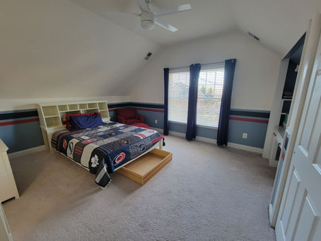 Attic bedroom with a bed, sports-themed quilt, red chair, bookshelf, and large window with navy curtains. Calm, tidy, and well-lit space.