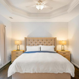 Bright bedroom with a large bed, beige headboard, white bedding, and two bedside lamps. Tray ceiling and large windows with curtains. Peaceful mood.