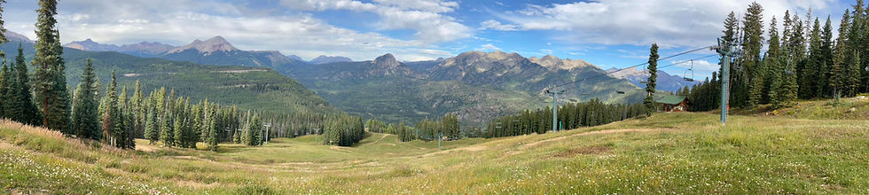 Wide mountain meadow with ski lifts, pine trees, and a small cabin under a partly cloudy sky and distant peaks