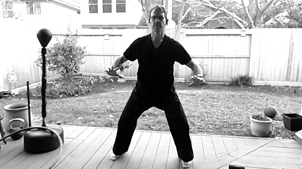 Man in black clothes practices martial arts on a wooden deck in a backyard. Fenced garden, potted plants, and training equipment visible.