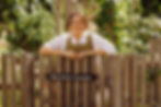Smiling woman in apron leans on a wooden fence in a lush garden, with a sign reading Kitchen Garden.