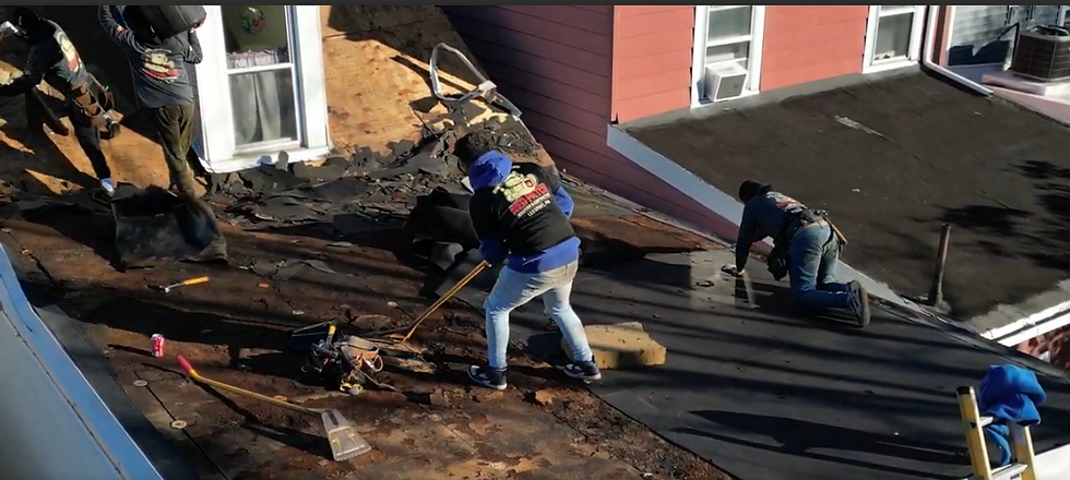 Workers in black jackets repairing a roof. One kneels, others use tools on asphalt shingles. Background shows red house and white windows.