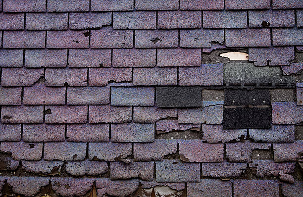 Weathered roof with missing and damaged dark shingles against a worn surface, showing signs of decay and neglect. No visible text.