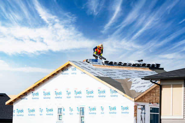 Worker in a yellow helmet installs shingles on a roof under a blue sky. The building is wrapped in Tyvek insulation material.