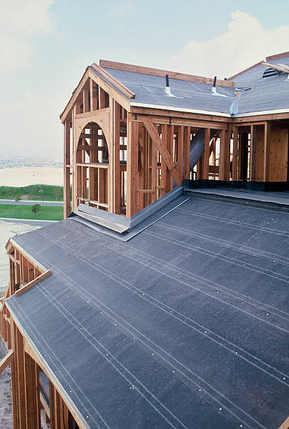 Wooden house under construction with exposed beams and black roofing, set against a cloudy sky and grassy landscape in the background.