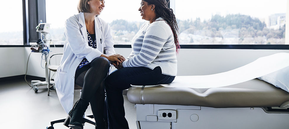 A doctor in a white coat smiles, holding hands with a patient in a striped sweater. They sit in a bright medical office, fostering a warm mood.