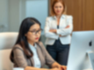 A woman sits at her computer while her boss stands with her arms crossed in the background