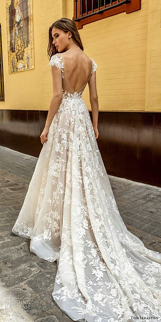 Bride wearing an A-line wedding gown featuring an open back, delicate shoulder sleeves, and a full lace skirt with a long train, standing outdoors on a cobblestone street.
