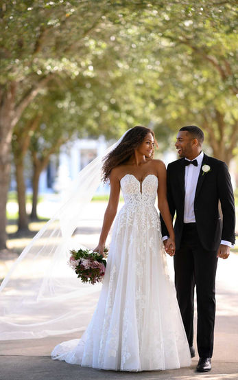 Bride wearing a strapless lace A-line wedding gown with a long veil, holding hands with the groom as they walk under a tree-lined path.