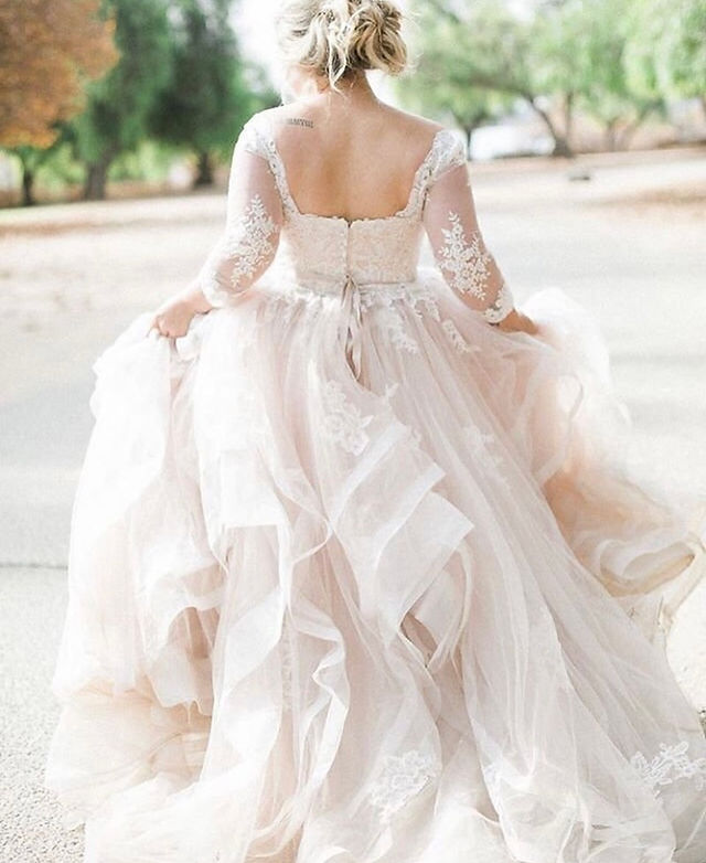 Bride captured from behind wearing a long-sleeve lace wedding gown with an open back and layered tulle skirt, walking outdoors on a sunlit path.