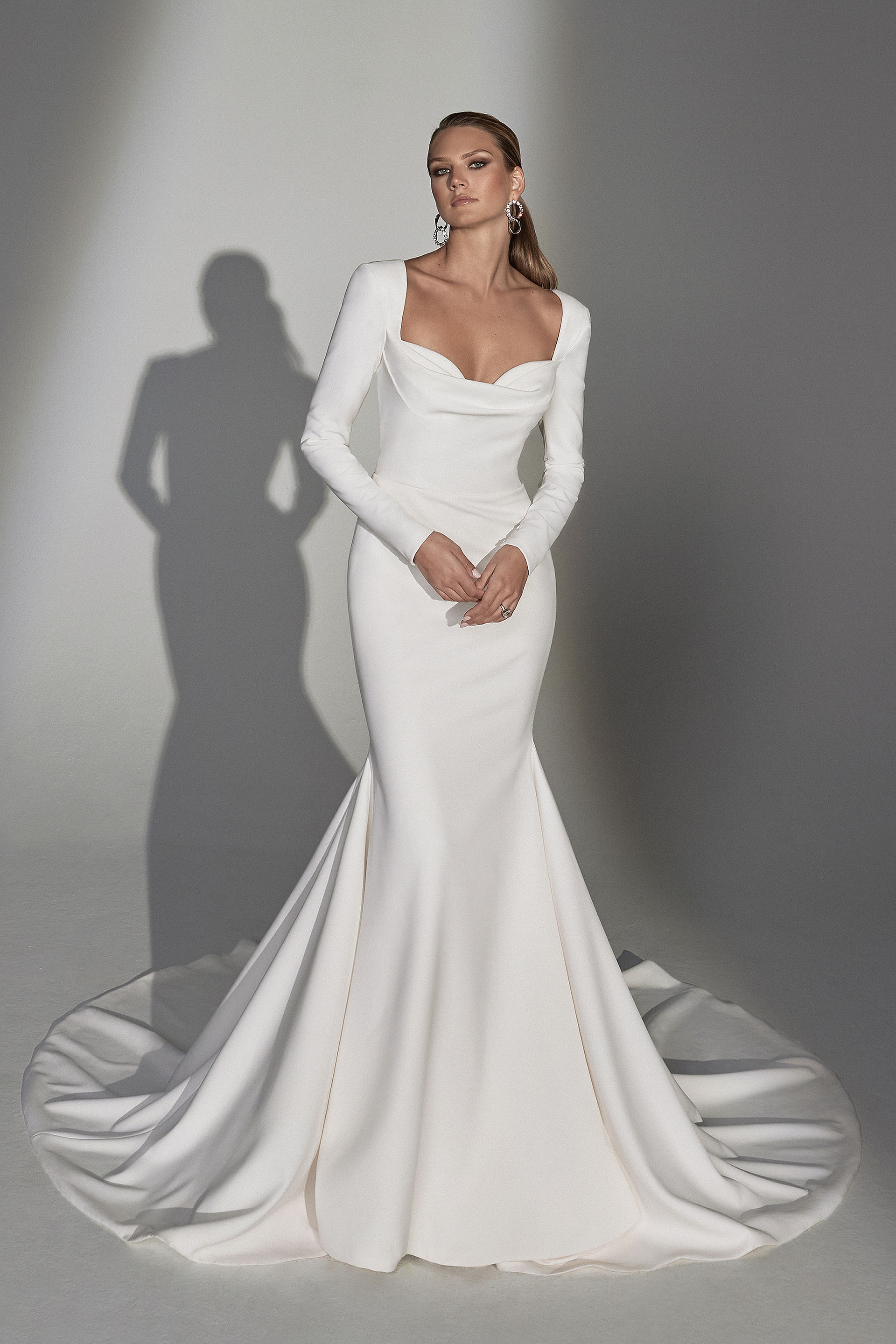 Bride wearing a long-sleeve satin fit-and-flare wedding dress with a structured square neckline and sweeping train in a studio setting