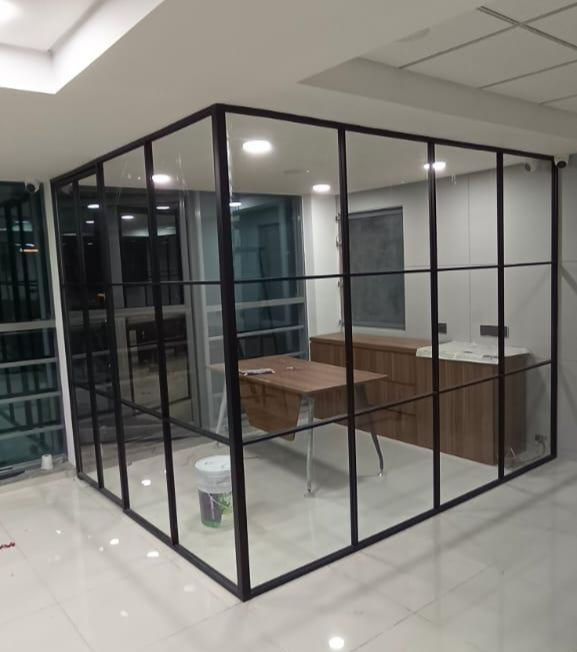 Glass-walled office with wooden desk, cabinets, and a paint bucket on the floor. Bright lighting and a modern, minimalist design.