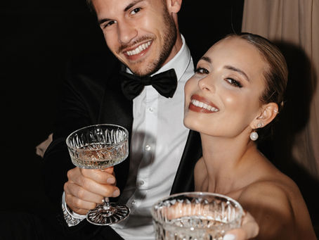 Fröhliches Paar in festlicher Abendkleidung stößt mit Kristallgläsern an. Beide lächeln in die Kamera – er trägt Smoking mit Fliege, sie elegante Ohrringe und glamouröses Make-up. Feierliche, stilvolle Atmosphäre.