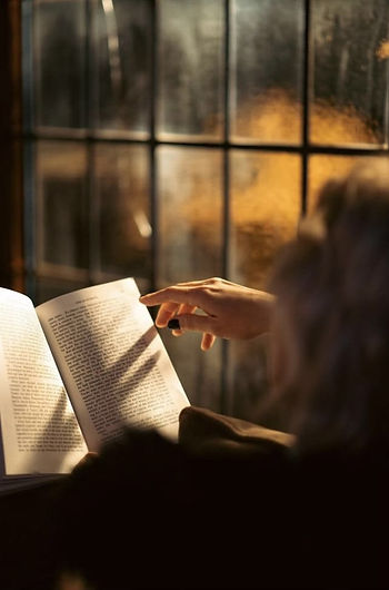 Bir kişi, loş bir ortamda açık bir kitap okuyor. Camın önünde, sıcak bir ışık içeriyi aydınlatıyor. Rahat ve huzurlu bir atmosfer.