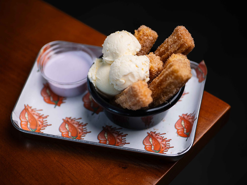 Churros and vanilla ice cream in a black bowl on a tray with orange lip designs, next to a small cup of purple sauce, on a wooden table.