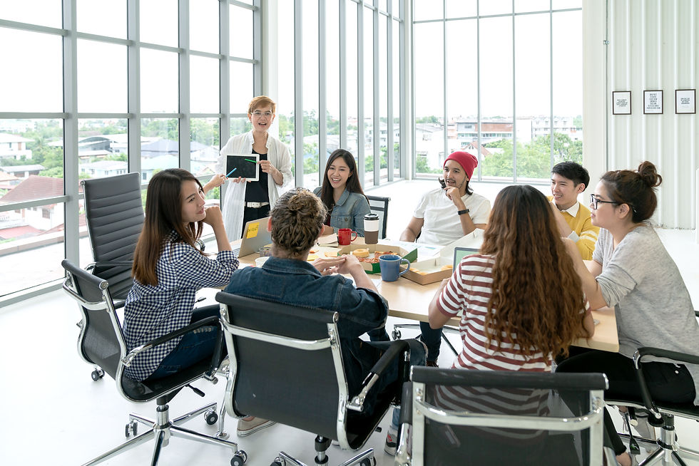Smiling coworkers in a bright glass office discuss around a table; a woman presents a tablet, with coffee and snacks nearby.