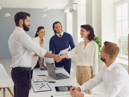 People in business attire shake hands in a bright office. Others smile, holding papers. A laptop and documents are on the table. They have found their content writer!