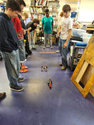 Students watch two small robots race across the classroom floor at school.