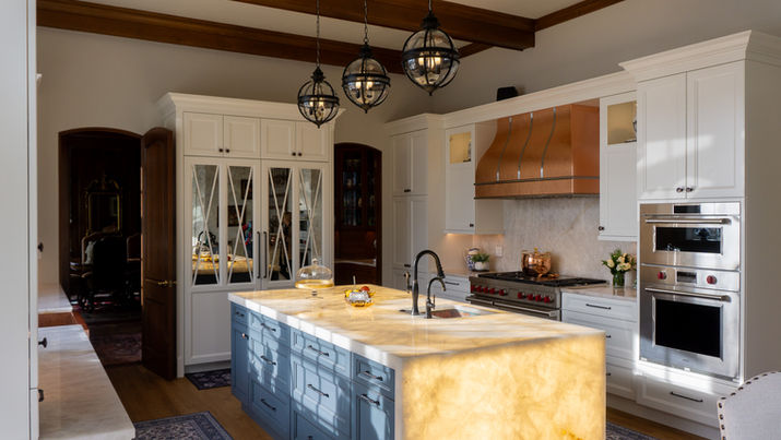 Luxury kitchen featuring a backlit Cristallo Traslux Ultra Quartzite island, full height quartzite splash, blue cabinetry, copper range hood, and warm ambient lighting highlighting the stone’s translucency.