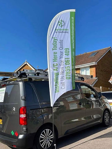 A van is parked on a street. A flag beside it advertises Hertfordshire Air Conditioning, offering installation services.