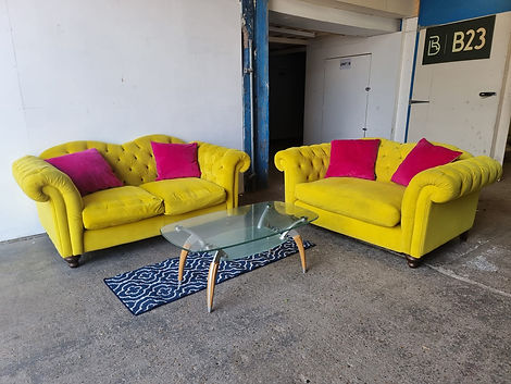 Two bright yellow, tufted sofas with magenta pillows sit in a room. A glass coffee table with wooden legs