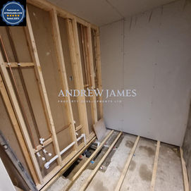 An unfinished room showing wooden framing, exposed plumbing, and a bare concrete floor