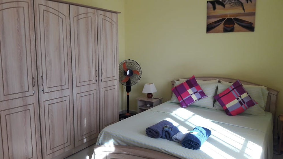Sunlit bedroom with a neatly made bed, pink plaid pillows, folded towels, wardrobe, fan, and beach wall art.
