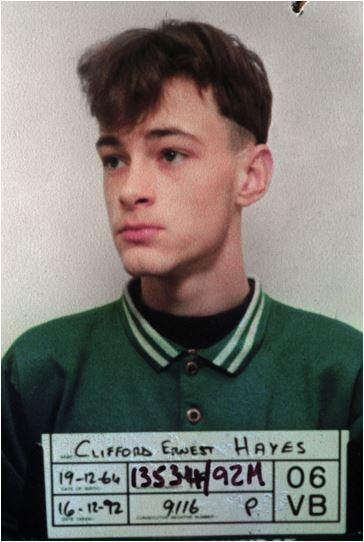 Young man in green shirt poses for mugshot holding a board labeled Clifford Ernest Hayes, looking serious.