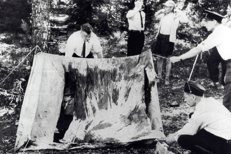 Investigators examine a damaged tent in a forest; three observers watch. The scene is tense, with trees visible in the background. Black and white photo.