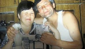 Smiling couple poses closely in a kitchen. The woman holds a glass. Warm, wooden cabinets form the background, creating a cozy atmosphere.