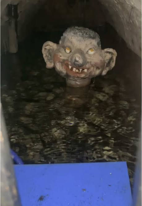 Creepy troll-like face floats in dark water inside a tunnel, with a blue platform in front.