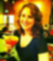 Woman with curly hair smiling, holding a cocktail with a lime slice in a warmly lit restaurant. Brown and orange background.