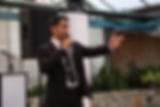 A professional great wedding MC engaging the crowd with a microphone at an outdoor wedding reception, surrounded by speakers and lush greenery.