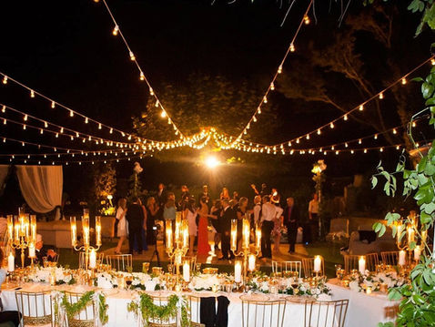 An outdoor luxury wedding reception at night with long tables, gold chairs, elegant candelabras, and guests dancing under a canopy of string lights.