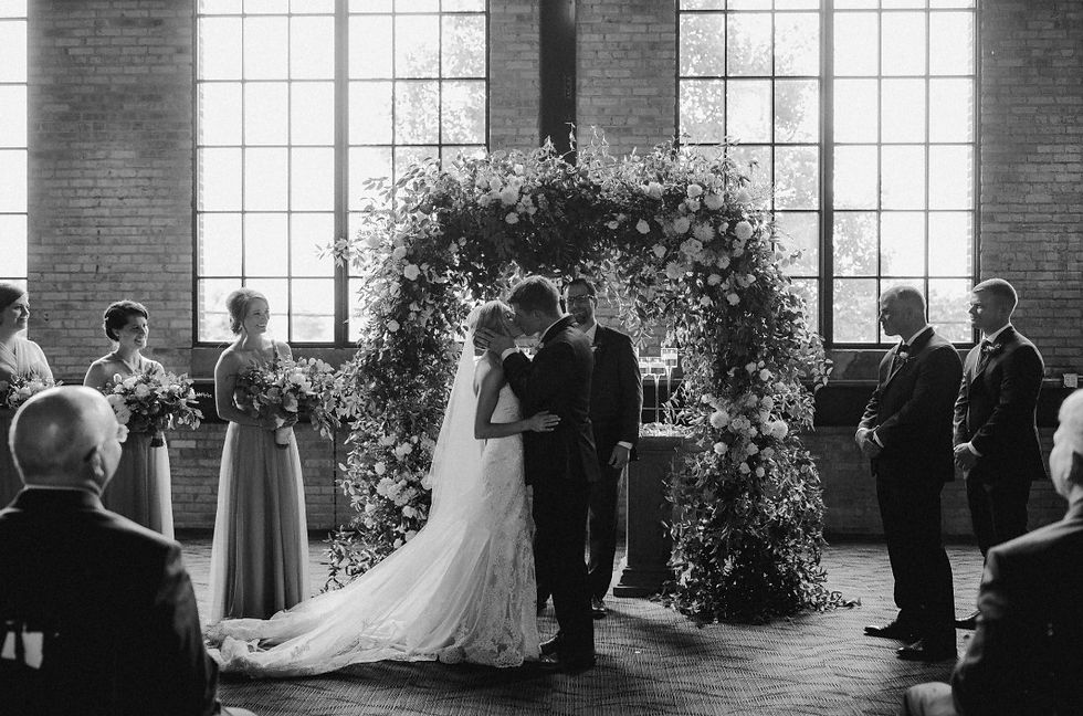 A bride and groom sharing their first kiss under a lush floral arch during a wedding ceremony in an industrial-style venue.