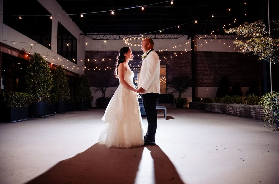 A bride and groom standing face-to-face in an urban courtyard at night, illuminated by a spotlight and overhead bistro string lights.