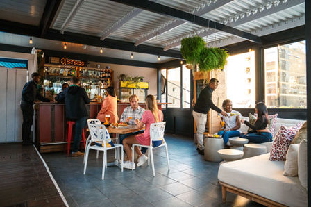 A lively and modern rooftop bar scene at The Grand Daddy Hotel, with patrons enjoying drinks and food at a counter bar, a dining table, and a comfortable lounge area with large windows overlooking the city.