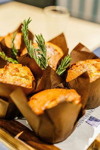 A close-up of a selection of golden-brown muffins in dark brown paper wrappers, presented in a wooden box with sprigs of fresh rosemary.