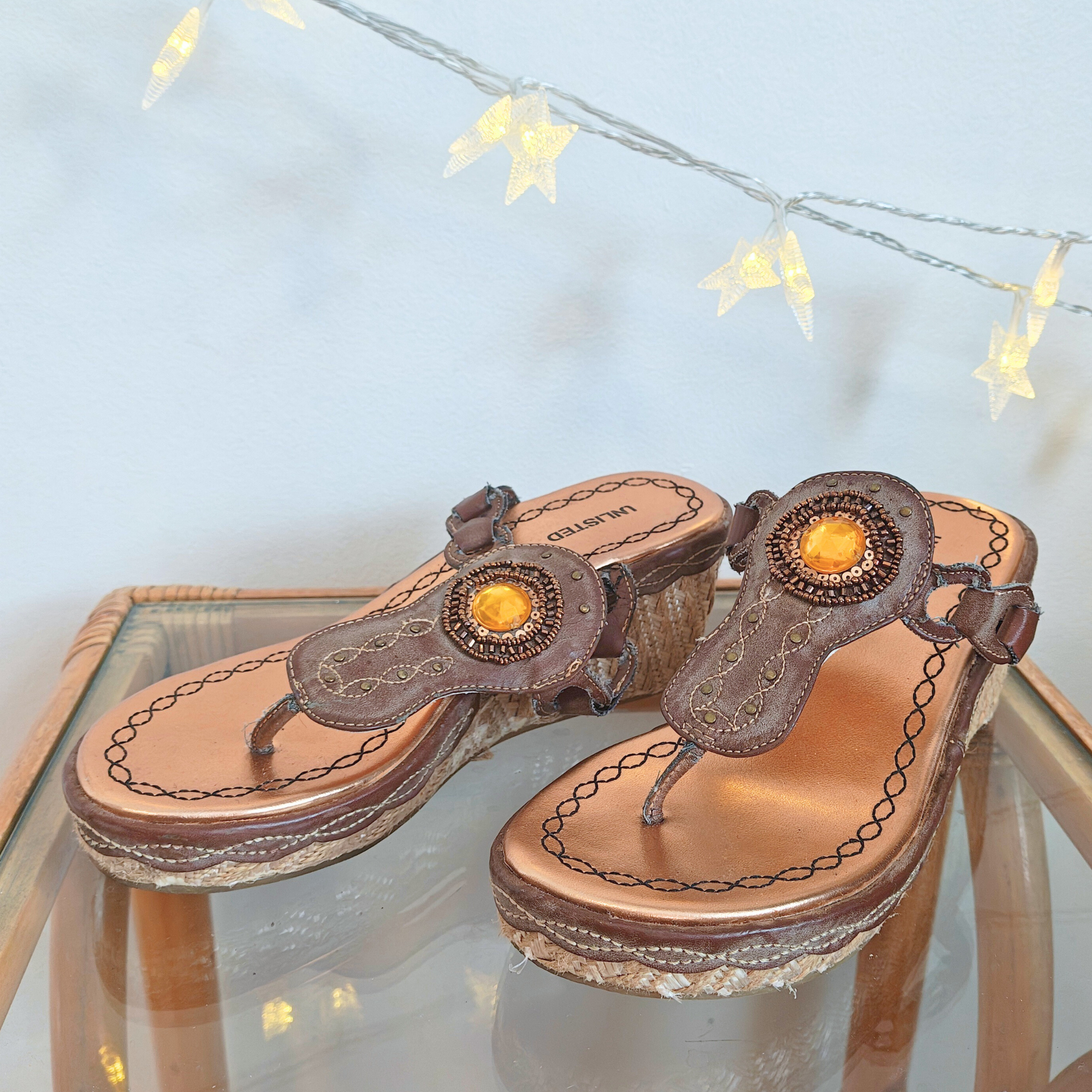 Brown sandals with orange embellishments on glass surface, festive lights background.