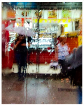 Visão altamente distorcida por água escorrendo no vidro, mostrando a silhueta de uma pessoa com guarda-chuva claro em frente a um comércio iluminado em amarelo e vermelho.