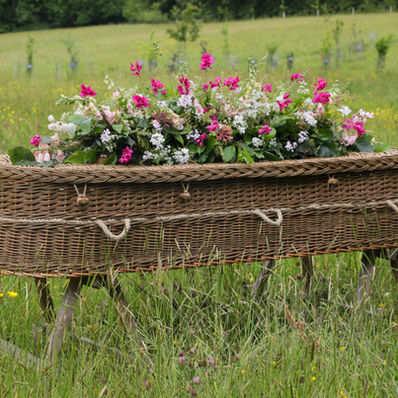 Hand‑woven willow coffin titled brown petal coffin in summer meadow - showcasing sustainable design and floral tribute
