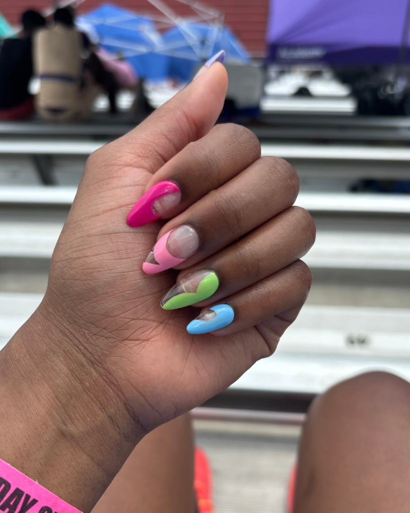 A close-up of a hand with colorful DIY nail art featuring pink, green, and blue nails, showcasing playful shapes and designs.