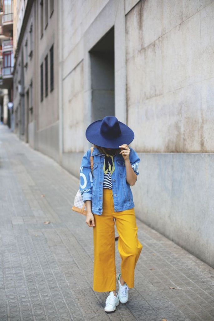 A fashionable person wearing a denim jacket, striped shirt, and mustard yellow wide-leg pants, standing on a street with a cobblestone pavement. They are wearing a navy blue hat and white sneakers, holding a bag while partially obscuring their face.