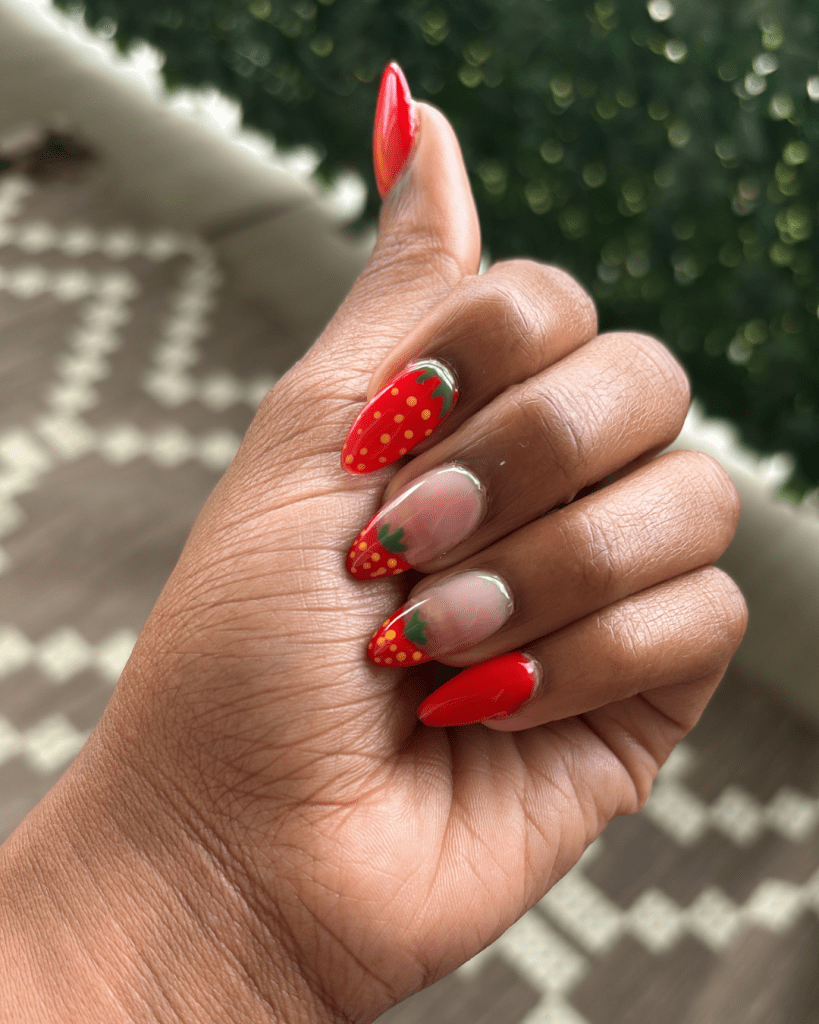 Close-up of hands showing DIY strawberry nail art, featuring red polish with green tips and yellow dots representing seeds.