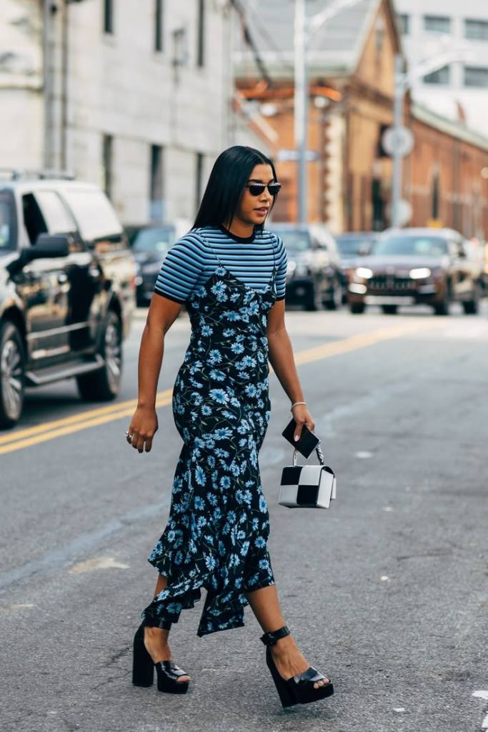 Woman walking on a city street, wearing a striped shirt layered over a floral dress, accessorized with sunglasses and carrying a small purse.