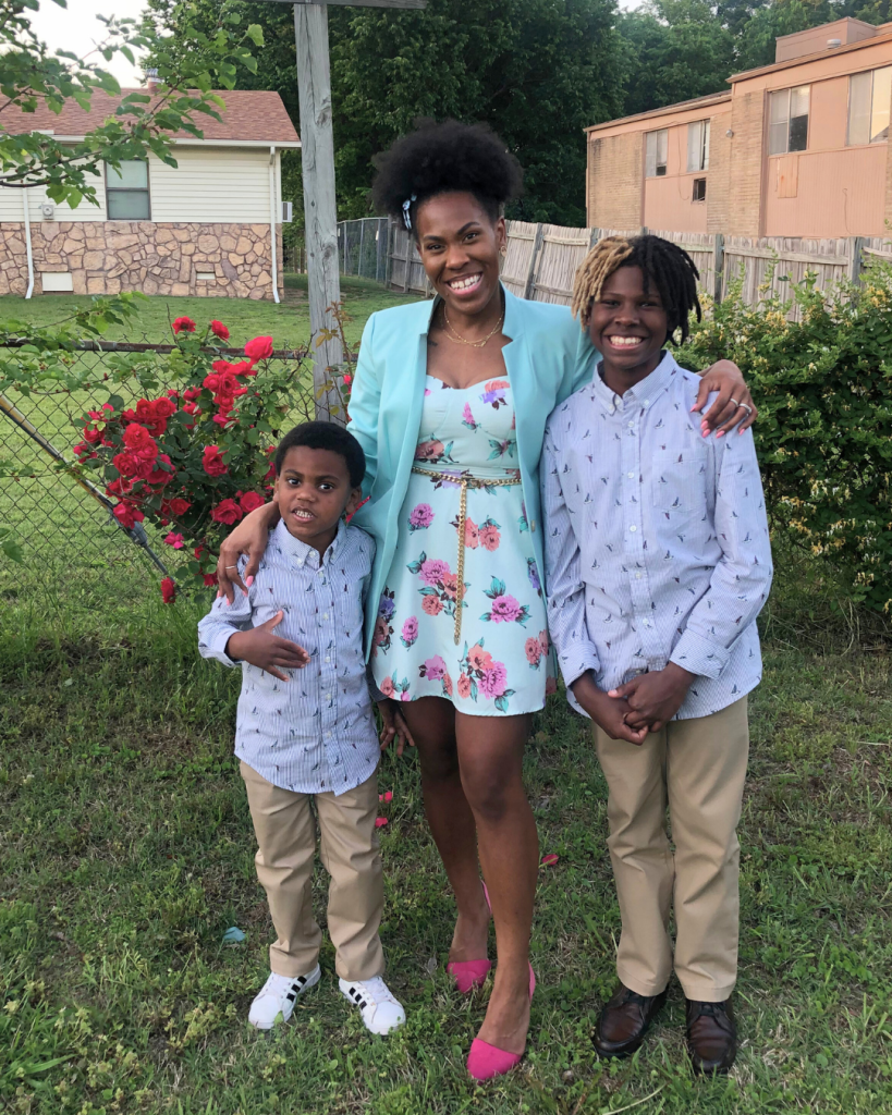 A mother smiling and standing with her two children in a garden, surrounded by blooming flowers. The mother is wearing a floral dress and a light jacket, while the children are dressed in collared shirts and slacks.