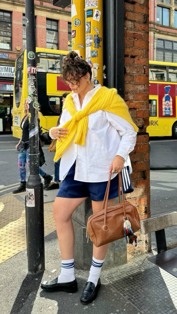 A person wearing a white shirt, a yellow knitted sweater draped around their shoulders, and navy shorts, standing near a brick wall with a lamppost covered in stickers. They hold a brown handbag and wear white socks with blue stripes.