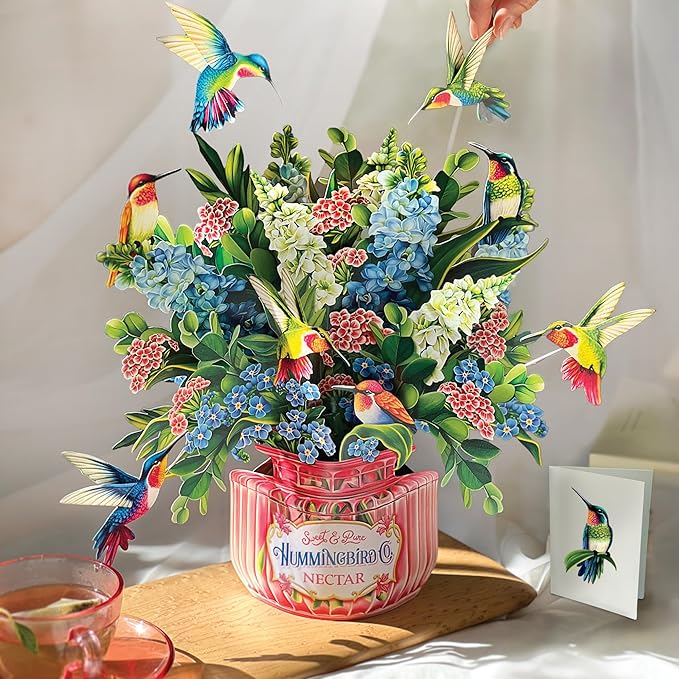 A vibrant pop-up paper bouquet featuring colorful flowers and hummingbirds, resting in a decorative container with the label 'Hummingbird Nectar'.